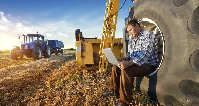 A farmer sat using a laptop. The farmer is sat beside farming machinery.