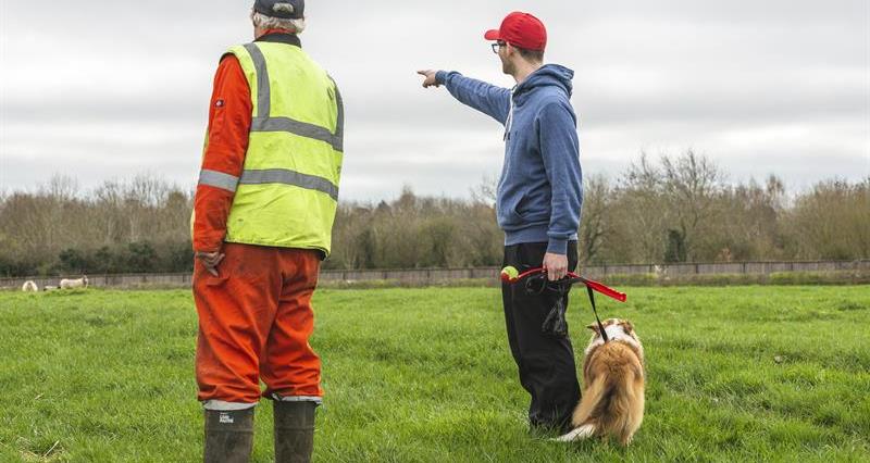 An image of a man walking a dog in a field talking to another man who is wearing a high vis vest