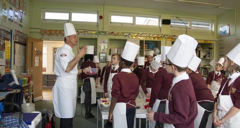 An image of primary school children in a classroom wearing aprons and chefs hats listing to a chef who is leading them in a practical lesson about food 