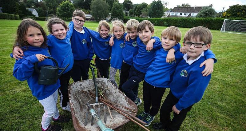 A group of year 2 and 3 pupils gathered around a wheelbarrow containing gardening implements, with the school field in the background