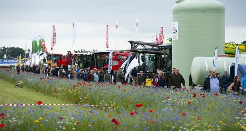 A picture of a group of people looking at agricultural machinery next to a grassy area containing lots of wildflowers