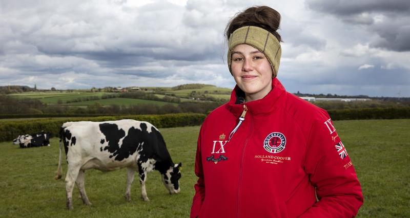 A photo of Jess Langton in a red jacket standing in a dairy cow field.