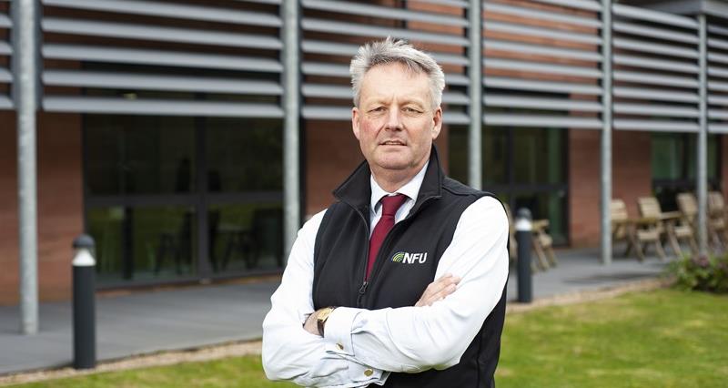NFU Vice President David Exwood stands with his arms foldedon some grass in front of NFU HQ. He wears a white shirt and red tie, and an NFU gilet.