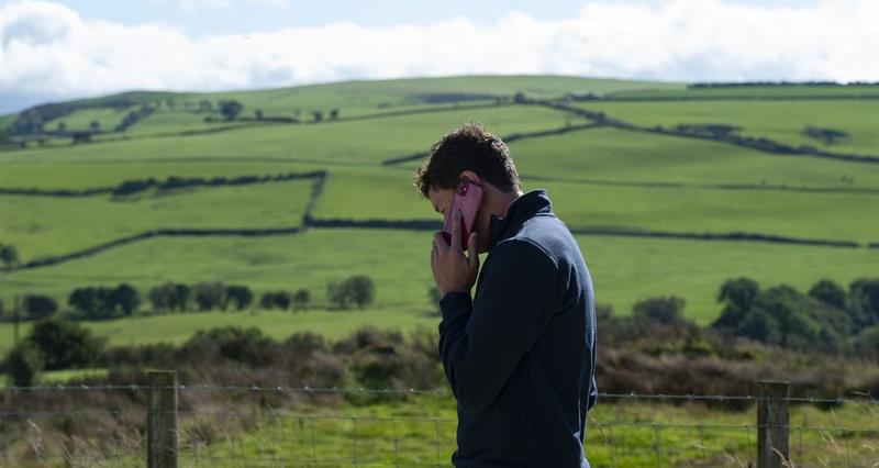 Man with a phone held to his ear standing next to a wire fence, with green fields behind him.
