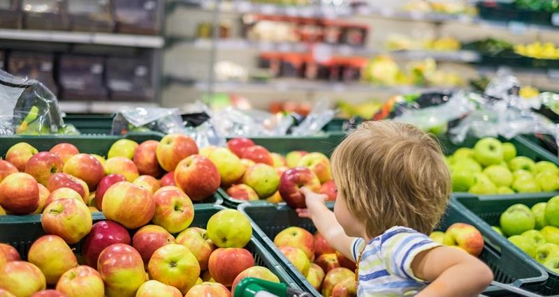 An image of a small boy sitting in a supermarket trolley reaching for an apple
