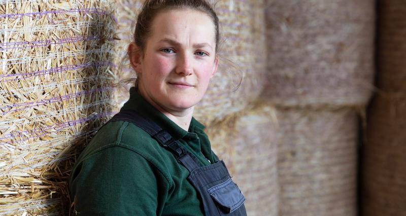 A photo of Darcy Johnson. She is stood sideways against a stack of hay bales.