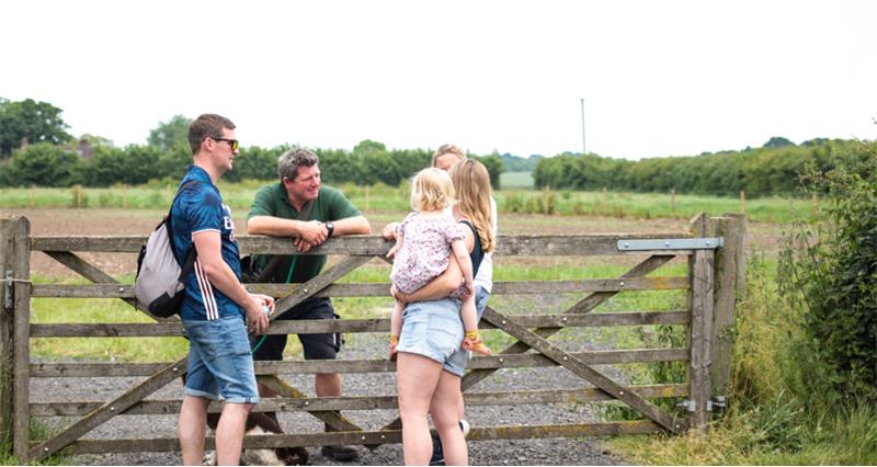 An image of a farmer stood behind a gate to a field and talking to a young family at Open Farm Sunday 2023. 