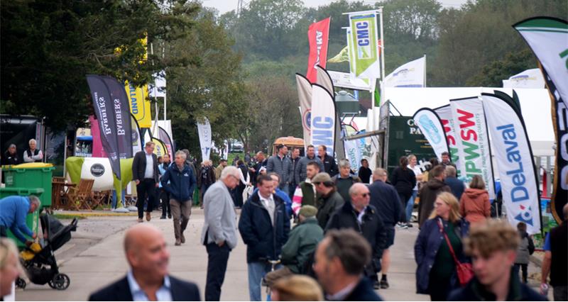 People walking at an agricuktural show, with flags and stalls visible along the sides of the road
