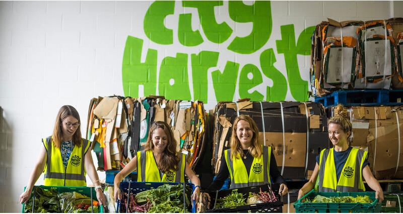 Four people from City Harvest stood in front of of a city harvest sign holding pallets of vege