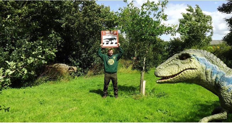 An image of Richard Bower holding up a dinosaur sign on his farm stood behind a dinosaur statue