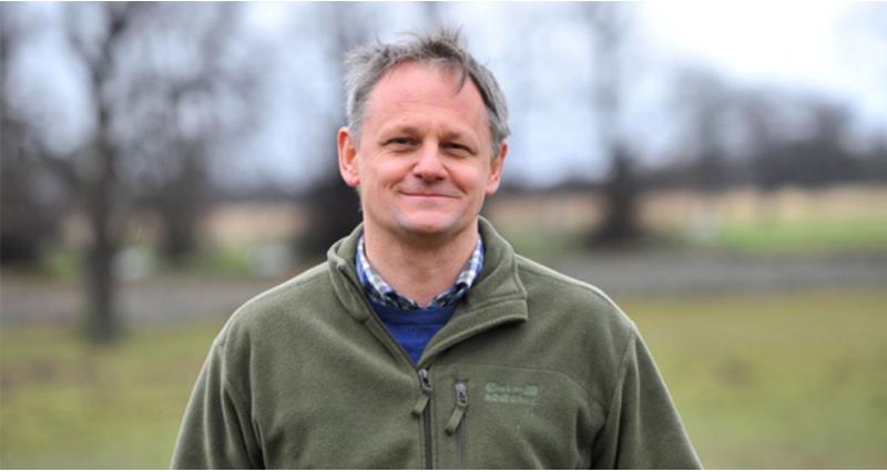 A portrait photo of farmer Andrew Blenkiron, who is smiling and wearing a green fleece, and is stood in front of a countryside landscape.