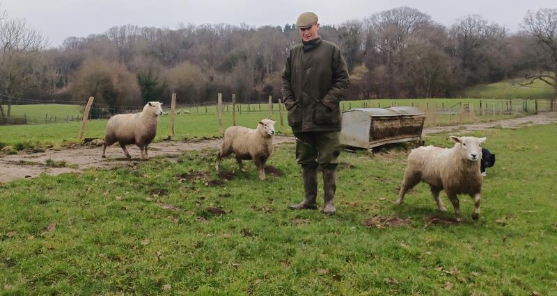 Kent farmer Tobin Bird standing in a field in a coat and wellington boots with some of his sheep